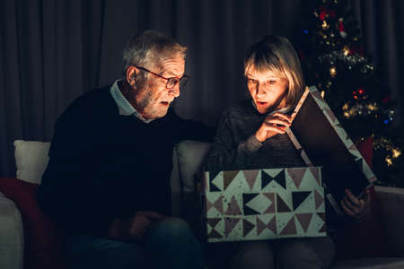 Senior man giving gift box to wife and having fun opening magic christmas box together sitting on...