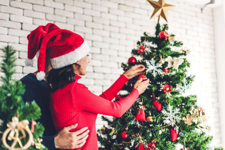 Romantic sweet couple in santa hats having fun decorating christmas tree and smiling while...