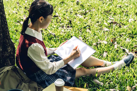 Woman relaxing with pen writing on a notebook sitting under a tree on grass in parkの写真素材