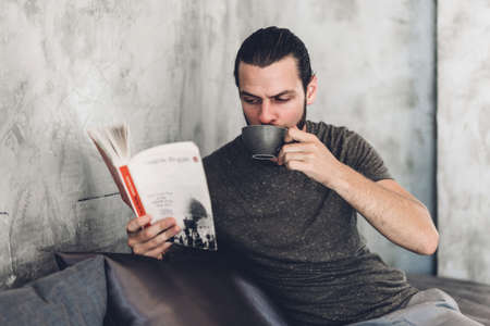 Handsome bearded hipster man relaxing holding and reading book with coffee while sitting on chair in cafeの写真素材