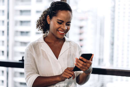 Smiling beautiful professional business african american black woman working and using smartphone standing in officeの写真素材