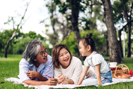 Portrait of happy grandfather with grandmother and little cute girl enjoy relax reading a book in summer park.Young girl with their laughing grandparents smiling together.Family and togethernessの写真素材