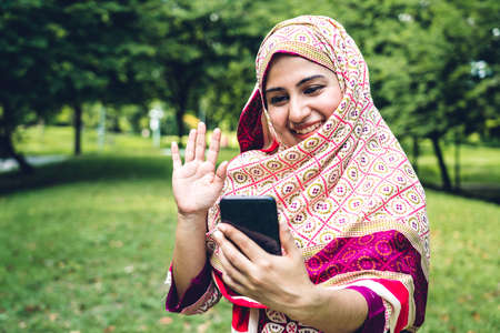 Portrait of happy arabic muslim woman with hijab dress smiling using and talking on the smartphone in summer parkの写真素材