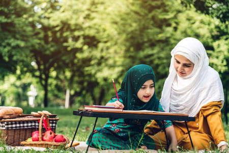 Portrait of happy muslim mother and little muslim girls child learning together with hijab dress smiling and enjoy relax reading and write a book in summer park.Education conceptの写真素材