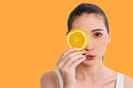 Portrait of beautiful joyful young pretty cute happy asian woman holding juicy orange fruit slice hiding eye behind and looking at camera isolated on orange background.Healthy food eating concept.Dietの写真素材
