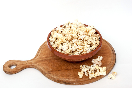 Wooden cutting board, clay bowl full of popcorn, some popcorn scattered.の写真素材