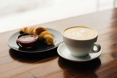 Cup of cappuccino with latte art and croissant with jam on the wooden table in cafe in the morningの写真素材
