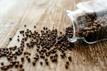 Black pepper is poured from a glass jar on a wooden surface