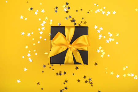 Black gift box with yellow bow on yellow background with sparkling confetti. Flat lay.の写真素材
