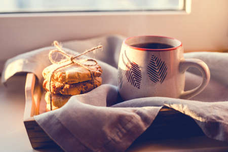 A steaming cup of tea or coffee with cookies by the window in the sunlight.