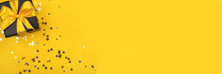 Black gift box with yellow bow on yellow background with sparkling confetti. Flat lay. Copy space.の写真素材