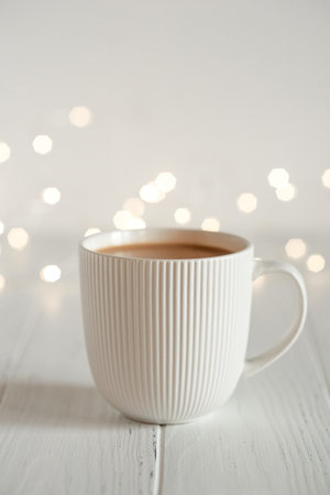 A white cup with hot coffee or tea on a white wooden table with Christmas lights.の写真素材