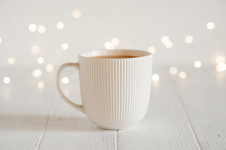 A white cup with hot coffee or tea on a white wooden table with Christmas lights.の写真素材