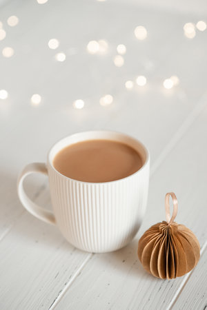 A white cup with hot coffee or tea on a white wooden table with Christmas lights.の写真素材