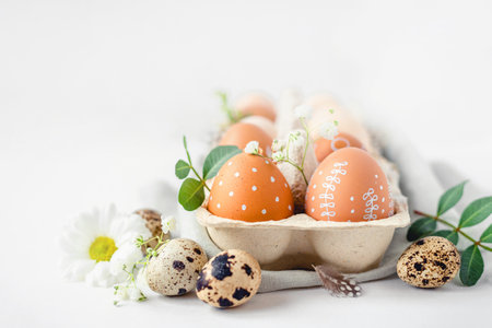 A delicate Easter background with hand-painted eggs in a box on a white table. Easter composition with flowers and feathersの写真素材