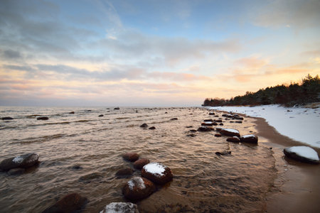 A view of the snow-covered Baltic sea coast at sunset. Stones in the water close-up. Coniferous forest in the background. Stunning cloudscape. Warm evening light. Kaltene, Latviaの写真素材