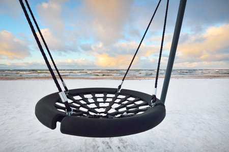 Modern swing on the snow-covered Baltic sea shore at sunset. Riga bay, Latvia. Colorful dramatic cloudscape. Deep cyclone, rough weather. Winter vacations, global warming themeの写真素材