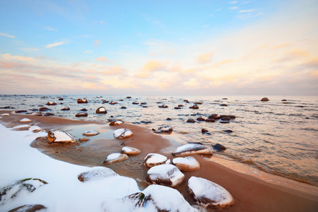 A view of the snow-covered Baltic sea coast at sunset. Stones in the water close-up. Clear blue sky with colorful clouds. Stunning cloudscape. Warm evening light. Idyllic winter scene. Kaltene, Latviaの写真素材
