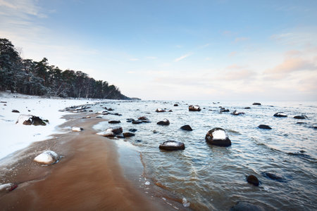 A view of the snow-covered Baltic sea coast at sunset. Stones in the water close-up. Coniferous forest in the background. Stunning cloudscape. Warm evening light. Kaltene, Latviaの写真素材