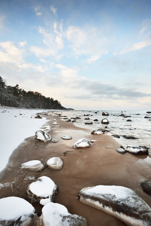 A view of the snow-covered Baltic sea coast at sunset. Stones in the water close-up. Coniferous forest in the background. Stunning cloudscape. Warm evening light. Kaltene, Latviaの写真素材