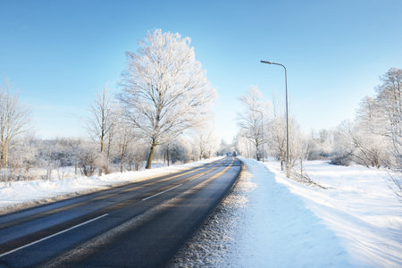 An empty asphalt road after cleaning. Street lanterns close-up. Car tracks in a fresh snow. Snow-covered birch forest in the background. Clear blue sky. Winter driving in Finland. Global warming themeの写真素材