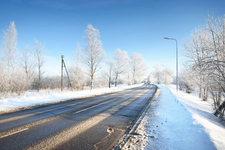 An empty asphalt road after cleaning. Street lanterns close-up. Car tracks in a fresh snow. Snow-covered birch forest in the background. Clear blue sky. Winter driving in Finland. Global warming themeの写真素材