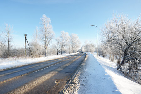 An empty asphalt road after cleaning. Street lanterns close-up. Car tracks in a fresh snow. Snow-covered birch forest in the background. Clear blue sky. Winter driving in Finland. Global warming themeの写真素材