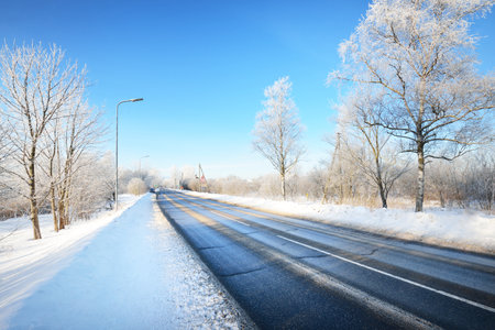 An empty asphalt road after cleaning. Street lanterns close-up. Car tracks in a fresh snow. Snow-covered birch forest in the background. Clear blue sky. Winter driving in Finland. Global warming themeの写真素材
