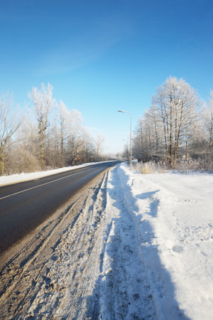 An empty asphalt road after cleaning. Street lanterns close-up. Car tracks in a fresh snow. Snow-covered birch forest in the background. Clear blue sky. Winter driving in Finland. Global warming themeの写真素材