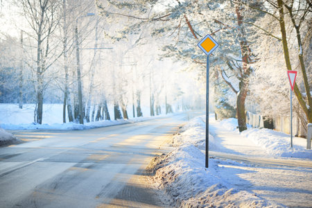 An empty snow-covered asphalt country road through the pine tree forest at sunset. Road sign close-up. Sunlight through the trees. Winter driving. Latviaの写真素材