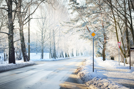 An empty snow-covered asphalt country road through the pine tree forest at sunset. Road sign close-up. Sunlight through the trees. Winter driving. Latviaの写真素材