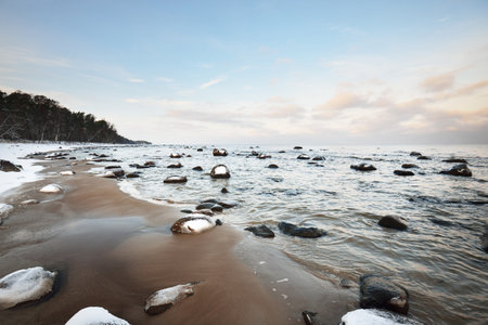 A view of the snow-covered Baltic sea coast at sunset. Stones in the water close-up. Coniferous forest in the background. Stunning cloudscape. Warm evening light. Kaltene, Latviaの写真素材