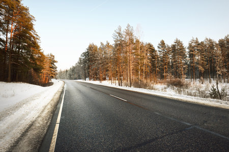 A clean empty asphalt country road through the coniferous forest at sunset. Pine and spruce trees after a blizzard. Winter driving. Latviaの写真素材