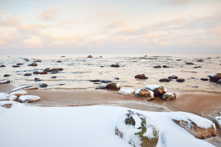A view of the snow-covered Baltic sea coast at sunset. Stones in the water close-up. Clear blue sky with colorful clouds. Stunning cloudscape. Warm evening light. Idyllic winter scene. Kaltene, Latviaの写真素材
