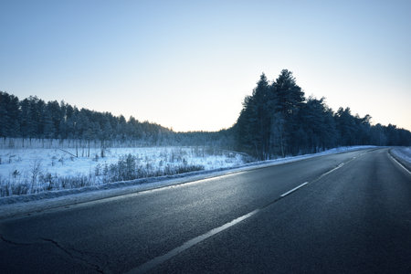 A clean empty asphalt country road through the coniferous forest at sunset. Pine and spruce trees after a blizzard. Winter driving. Latviaの写真素材