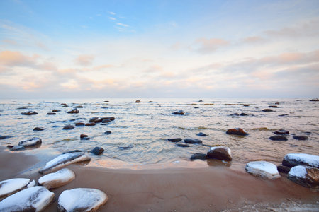 A view of the snow-covered Baltic sea coast at sunset. Stones in the water close-up. Clear blue sky with colorful clouds. Stunning cloudscape. Warm evening light. Idyllic winter scene. Kaltene, Latviaの写真素材