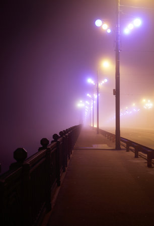 Pedestrian walkway through the illuminated empty Stone bridge in a thick fog at night. Lanterns close-up. Daugava river, Riga, Latvia. Concept image, neon colorsの写真素材