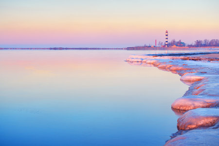 Frozen Baltic sea shore at sunset. Ice fragments and stones close-up. Lighthouse in the background. Colorful pink sky.Reflections on the water. Natural mirror. Latvia. Symbol of hope and peaceの写真素材