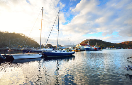 Motorboats, sailing and fishing boats moored to a pier in a small village in fjords. Hidra island, Rogaland region, Norway. Traditional architecture. Dramatic evening sky. Travel destinations themeの写真素材