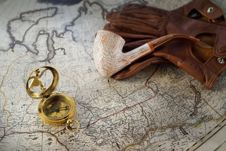 Retro style golden compass (sundial), elegant smoking pipe, brown leather gloves, old white nautical chart close-up. Vintage still life. Travel gentleman set. Planning travel, sailing, adventureの写真素材