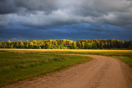 An empty country road with a sharp turn through the fields after the rain. Dramatic sky, epic cloudscape. Fickle weather. Latvia. Cyclone, storm, meteorology, ecology, climate changeの写真素材