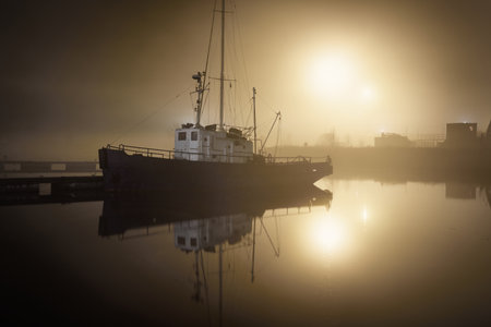 A fishing boat moored to a pier in a fog at night. Port cranes in the background. Yacht club...