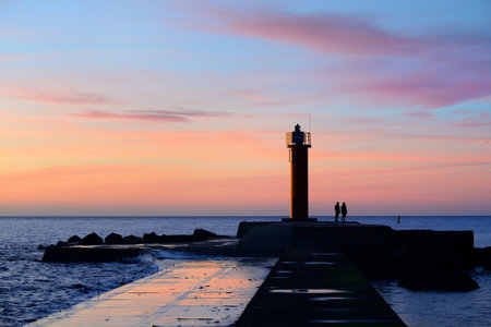 A couple of people standing near the orange lighthouse. Colorful sunset sky. Stunning cloudscape. Long exposure. National landmark, sightseeing, recreation theme. Baltic sea, Riga bay, Latviaの写真素材