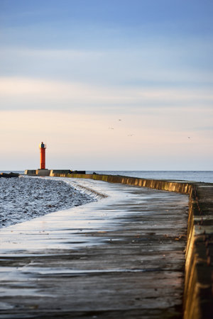An empty promenade to the orange lighthouse with solar battery against colorful sunset sky. Stunning cloudscape. Long exposure. Environmental conservation theme. Baltic sea, Riga bay, Latviaの写真素材