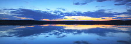 Cloudy blue sky with glowing golden sunset clouds above the forest lake. Symmetry reflections on the...