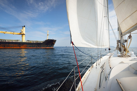 Large black cargo crane ship anchored in Strait of Gibraltar. A view from the yacht, white sails close-up. Summer Atlantic sailing near Spain and Africa. Clear blue skyの写真素材