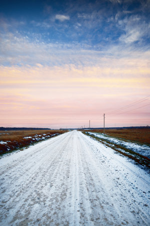 Snow-covered country road through the fields, transformer poles close-up. Forest in the background. Dramatic sunset sky with colorful clouds. Winter landscape. Latviaの写真素材