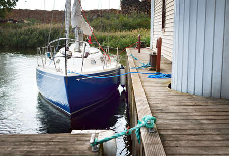Blue sloop rigged yacht moored to a wooden pier in a small village, close-up. Firewood in the...