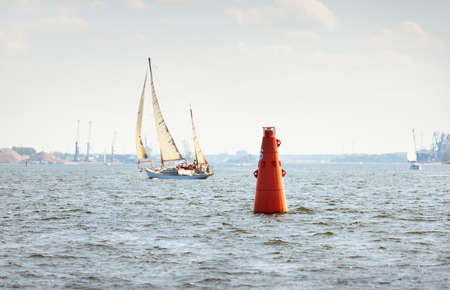 Red navigational buoy (a port mark) close-up. Old vintage wooden sailboat (yawl) close-up, sailing...