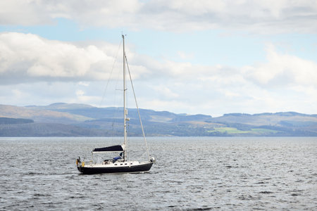 Black sloop rigged yacht sailing in Crinan canal on a cloudy day, close-up. Rocky shores in the background. Dramatic blue sky. Ardrishaig, Scotland, UK. Sport and recreation themeの写真素材